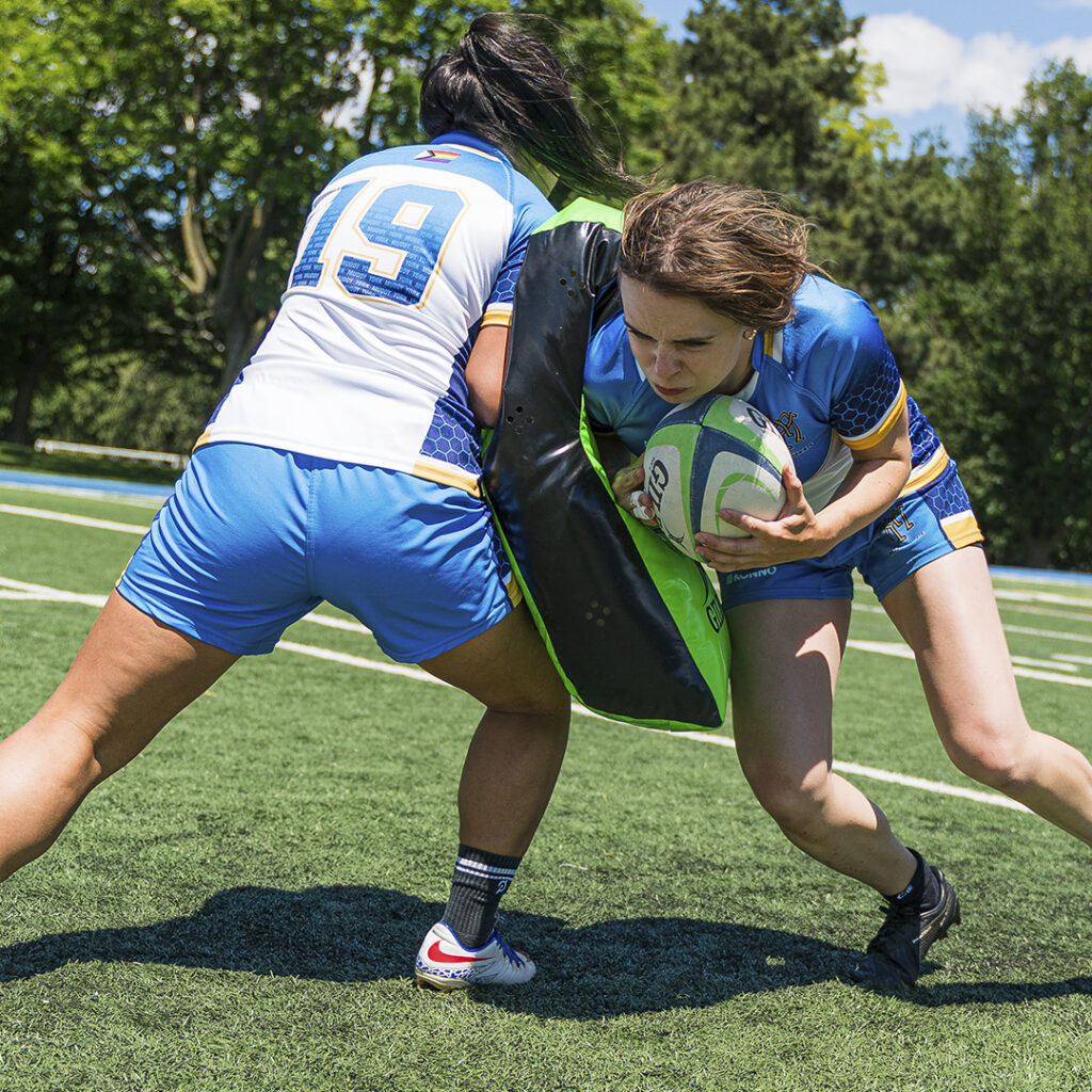A Muddy York team member carrying the ball into contact against her teammate, holding a ruck shield.